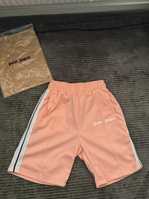 Palm Angels rosa shorts med vita ränder - Säljer ett par snygga rosa shorts från Palm Angels med vita och svarta ränder längs sidorna. Shortsen har elastisk midja och fickor på båda sidor. Ikonisk Palm Angels-logga framtill. Perfekta för sommaren och riktigt sköna att bära. Helt oanvända för de för för stora