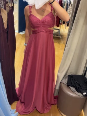 Lång rosa balklänning med volangband - Säljer en elegant långklänning i rosa med volangprydda axelband och v-ringad överdel. Klänningen har en markerad midja, lätt utställd kjol och fina draperingar på bysten. Materialet känns mjukt och faller snyggt.