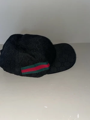 Svart keps från Gucci med GG-mönster - Svart keps från Gucci med klassiskt GG-monogram över hela kepsen. Snygg detalj med grön och röd rand på sidan. Böjd skärm och justerbar passform. Materialet är canvas med exklusiv känsla, perfekt för dig som gillar streetstyle och lyx.