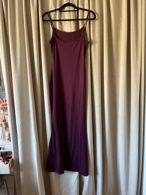 Vinröd långklänning med smala axelband - Vinröd långklänning med smala axelband och enkel, rak siluett. Tillverkad i mjuk polyester med lite stretch för en bekväm passform. Perfekt för dig som gillar minimalistisk stil och vill ha en klänning som känns både trendig och tidlös.