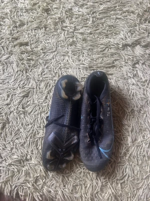 Nike Mercurial svarta fotbollsskor - Säljer ett par svarta Nike Mercurial fotbollsskor med blå detaljer och snörning. Skorna har låg profil och mönstrad ovandel för bra bollkontroll. Utrustade med fasta dobbar för spel på gräs. Perfekta för dig som vill ha snabba och lätta skor på planen.