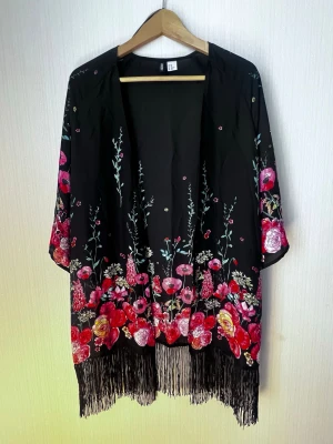 Svart kimono med blommigt mönster och fransar - Snygg svart kimono från H&M med färgstarkt blommigt mönster i rosa, rött och lila. Modellen har vida ärmar och långa fransar längs nederkanten. Perfekt för en chill dag hemma eller som statement-plagg i din loungewear-garderob. #h&m #kimono