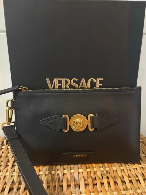 Versace Svart kuvertväska - Stilren svart kuvertväska från Versace i äkta kalvläder med guldfärgade detaljer och ikonisk Medusa-logga framtill. Väskan har avtagbar handledsrem och dragkedja upptill. Perfekt för dig som vill ha en lyxig accessoar med statement-känsla. Kvitto, låda och dustbag medföljer!