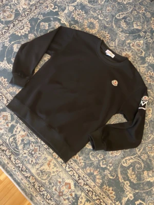 Svart sweatshirt från Moncler - Svart sweatshirt från Moncler med rund halsringning och klassiska Moncler-loggor på bröstet och ena ärmen. Tröjan har en relaxed fit och ribbade muddar vid ärmslut och nederkant. Perfekt för en clean och stilren look.