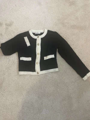 Svart kofta med vita detaljer - Snygg svart kofta med vita ribbade kanter och guldknappar framtill. Koftan har två dekorativa fickor. Perfekt för att lyfta en enkel outfit med en klassisk touch. I stl 158/164 men passar även xs/xxs