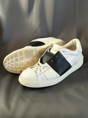 Valentino vita sneakers med svart detalj - Snygga vita sneakers från Valentino med en bred svart läderdetalj över snörningen. Skorna har klassisk låg profil, rund tå och platt sula med nitar baktill. Perfekta för dig som gillar stilrena och exklusiva sneakers med en twist.
