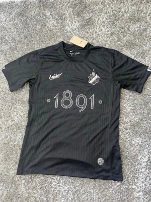 Svart AIK fotbollströja Nike - Hej kära gnagare. Snygg svart AIK fotbollströja från Nike med Dri-FIT-material. Tröjan har AIK:s klubbmärke, årtalet 1891 tryckt på bröstet och rund hals. Perfekt för träning eller match och har en sportig, modern look.