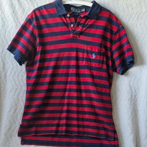 Randig pikétröja från Ralph Lauren - Klassisk pikétröja från Ralph Lauren i marinblått och rött med horisontella ränder. Tröjan har korta ärmar, krage, knappar vid halsen och en liten broderad logga på bröstet. Perfekt för en avslappnad och stilren look.