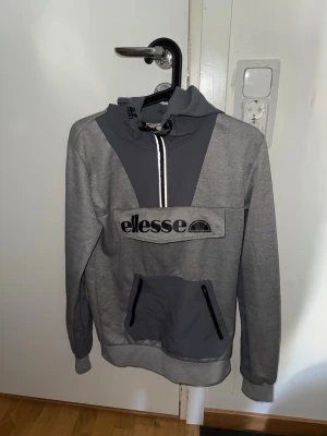 Grå hoodie från Ellesse med dragkedja - Snygg grå hoodie från Ellesse med stor logga framtill, dragkedja vid halsen och praktisk magficka med svarta detaljer. Hoodien har en kontrastfärgad huva och muddar vid ärmslut och nederkant. Perfekt för en avslappnad streetwear-look.