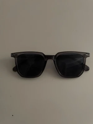Gråa solglasögon - Snygga solglasögon med transparent grå båge och mörka fyrkantiga glas. Klassisk och stilren design som passar till de flesta outfits. Perfekta för soliga dagar och ger en cool vibe.