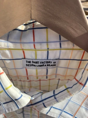 Vit rutig skjorta från The Shirt Factory - Vit skjorta från The Shirt Factory, designad av Linnéa Braun. Skjortan har ett färgglatt rutmönster i blått, rött, gult och orange. Klassisk krage, knäppning framtill och lång ärm. Perfekt för dig som gillar stilrena men ändå lekfulla plagg. Ingen doft från ärmhåla, 