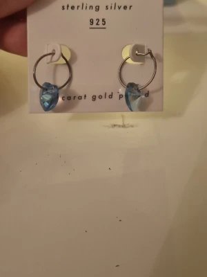 Silverörhängen med blå hjärthänge - Snygga örhängen i sterling silver 925 med runda hoops och blå hjärtformade hängen i glas eller kristall. Perfekta för att ge en färgklick till din stil. Enkla att bära och passar till många olika outfits.