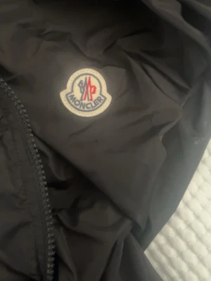 Svart vindjacka från Moncler - Snygg svart vindjacka från Moncler med klassisk logotyp på bröstet och innerfoder med märkets emblem. Jackan har dragkedja och tryckknappar framtill, perfekt för en stilren och sportig look.