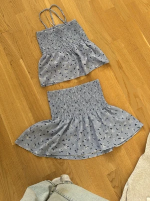 Blått blommigt set med topp och kjol - Säljer ett sött tvådelat set i ljusblått med små vita och svarta blommor. Setet består av en smockad topp med tunna axelband och en matchande smockad kjol med volang. Perfekt för en somrig look!