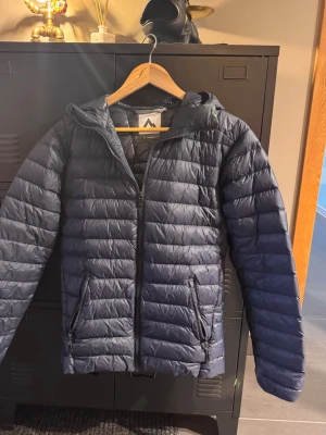Mörkblå dunjacka med huva - Snygg mörkblå dunjacka från Mckinley med huva och dragkedja framtill. Jackan har två framfickor med dragkedja och quiltad design som ger en sportig look. En perfekt overall jacka. Storleken ligger på herr XS. Ord pris 1000kr