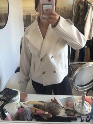 Vit ullkappa med bred krage - Stilren vit kappa med dubbelknäppning och bred krage. Kortare modell som ger en modern look, perfekt att matcha med jeans eller kjol. Kappan har stora knappar framtill och är tillverkad i ett ull-liknande material för extra värme.