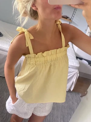 Gul linnetopp med knytband - Söt ljusgul linnetopp med knytband på axlarna och rynkad kant upptill. Toppen är ärmlös och har en luftig, loose passform som passar perfekt till varma dagar.