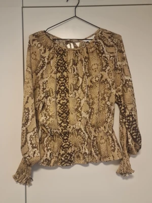 Beige blus med ormmönster - Trendig beige blus med ormmönster och långa ärmar. Blusen har resår och volangdetaljer vid ärmslut och midja samt en liten knytning i nacken. Tillverkad i ett lätt och luftigt material som ger en snygg passform.