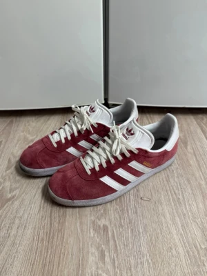 Adidas Gazelle vinröda sneakers - Snygga Adidas Gazelle sneakers i vinrött mocka med vita detaljer och klassiska tre ränder på sidorna. Skorna har vit plös och snörning samt gummisula. Skriv för fler frågor💪🏼