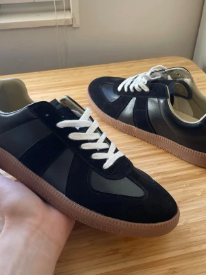 Svarta maison margiela - Stilrena svarta sneakers med paneler i mocka och släta partier i läder. Skorna har vita snören och en klassisk gummisula i brunt. Perfekta för dig som gillar en clean och tidlös look. Låg modell med bekväm passform.