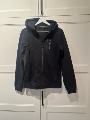 Svart hoodie från sail racing  - Svart hoodie med dragkedja från sail racing. Klassisk modell med huva, två sidofickor och logga på bröstet samt en detalj på huvan. Mjuk bomullsblandning och ribbade muddar vid ärmslut och nederkant. Perfekt för en avslappnad stil. Nypris (999)