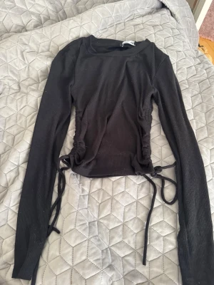 Svart ribbad topp med dragsko - Trendig svart långärmad topp med ribbad struktur och dragsko i sidorna som ger en snygg rynkad effekt. Tight passform och rund halsringning. Perfekt att styla med jeans eller kjol för en cool look.