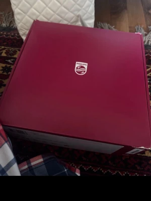 Philips laser aparat med sladd - Elektroniktillbehör från Philips i originalförpackning, levereras med vit strömadapter och kabel. Förpackningen är vinröd med Philips-logga. Perfekt för dig som gillar att lasra dig hemma och vill ha något från ett välkänt märke.
