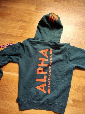 Grön hoodie från Alpha Industries - Snygg grön hoodie från Alpha Industries med stor orange logga på ryggen och huvan. Hoodien har dragkedjeficka på ärmen med röd tag och klassisk känguruficka framtill. Mjuk bomullskänsla och justerbar huva med snören.