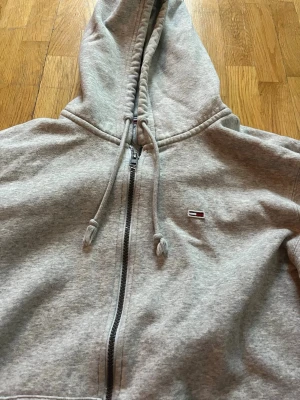 Grå hoodie från Tommy Hilfiger - Snygg grå hoodie från Tommy Hilfiger med dragkedja framtill, justerbar huva med snören och klassisk logga på bröstet. Tillverkad i mjuk bomullsmix och har två stora fickor framtill. Perfekt för chill dagar eller när du vill ha en clean look.