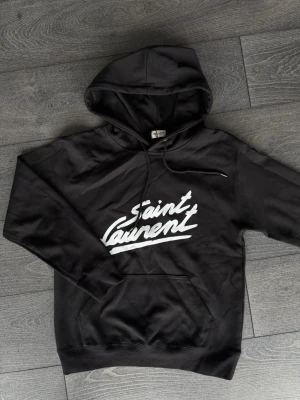 Svart Saint Laurent hoodie - Svart hoodie från Saint Laurent med vit logotyp i graffiti-stil på bröstet. Klassisk modell med huva, dragsko och känguruficka framtill. Perfekt för en cool och avslappnad streetwear-look.