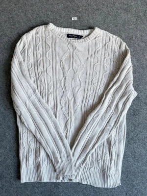Ljusgrå kabelstickad tröja Nautica - Kabelstickad tröja från Nautica i ljusgrå färg. Tröjan har rund halsringning, ribbade muddar och ett klassiskt kabelmönster framtill. Perfekt för dig som gillar en stilren och tidlös look.