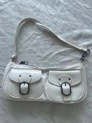 Vit axelremsväska med spännen - Snygg vit handväska i rektangulär form med två framfickor som har dekorativa spännen i silverfärg. Väskan har en kort axelrem och stängs med dragkedja upptill. Perfekt accessoar för en trendig look.🤍 pris kan diskuteras 