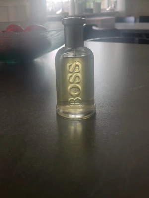Hugo Boss parfymflaska - Säljer en stilren parfym från Hugo Boss. Flaskan är genomskinlig med silverfärgad kork och har BOSS ingraverat på framsidan. Parfymen har en ljusgul nyans och flaskan är i glas med rundad form. Jag kan diskutera pris