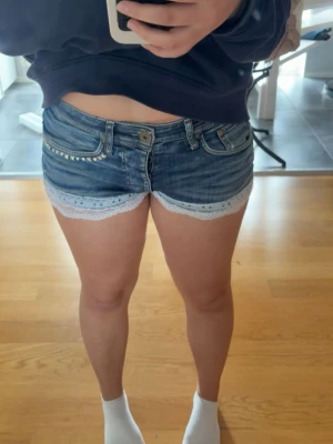 Blå jeansshorts med spetskant - Snygga blå jeansshorts med låg midja och vit spetskant längst ner. Shortsen har inga fickor baktill, bälteshällor och dekorativa nitar vid ena fickan. Perfekta för sommaren och ger en fräsch och trendig look.