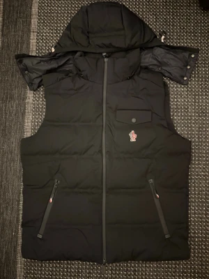 Moncler väst - Stilren svart dunväst från Moncler med avtagbar huva, två dragkedjefickor och en bröstficka med knapp. Västen har Moncler-logga på bröstet och är tillverkad i ett glansigt material som känns lyxigt. Perfekt för lager-på-lager och streetwear.