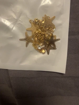 Guldfärgat fotarmband/armband med sjöstjärnor - Snyggt guldfärgat armband med små sjöstjärneberlocker och glittrande stenar. Kedjan är tunn och smidig, perfekt för att ge en trendig touch till din stil. Armbandet har flera detaljerade sjöstjärnor i metall och gnistrande pärlliknande dekorationer.