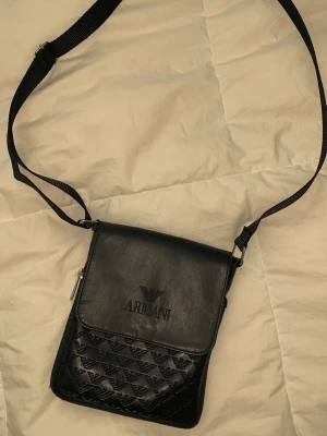 Svart axelväska från Armani i äkta läder  - Snygg svart axelväska från Armani med justerbar rem. Väskan har 3 stycken fickor med präglad logotyp och mönster av små Armani loggor på framsidan. Tillverkad i äkta läder. Perfekt för att bära runt dina dagliga redskap på ett enkelt och effektivt sätt. Köpt i Italien för ungefär 3 år sedan, så kvittot har jag lyckats slarva bort. !Skriv vid intresse så kommer vi överens om ett tillfredsställande pris tillsammans! Mvh.
