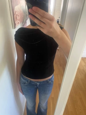 Svart sammets topp från Gina Tricot - Snygg svart topp i sammetsmaterial från Gina Tricot. Toppen har en croppad passform och är ärmlös med en draperad effekt vid axlarna. Perfekt att styla med jeans eller kjol för en trendig look.