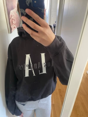 Svart hoodie med LA-tryck Ginatricot  - Cool svart hoodie med stor vit text 'LA' och grå text 'Los Angeles' på bröstet. Klassisk modell med huva och ribbade muddar. Perfekt för en avslappnad streetstyle-look. Materialet känns mjukt och skönt mot huden.