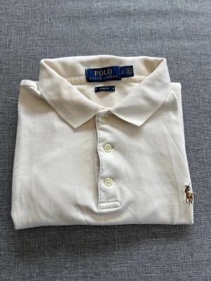 Vit piké från Polo Ralph Lauren - Klassisk vit pikétröja från Polo Ralph Lauren i slim fit-modell. Tröjan har krage, knäppning med tre knappar och den ikoniska broderade loggan på bröstet. Tillverkad i mjuk bomull för en bekväm känsla.