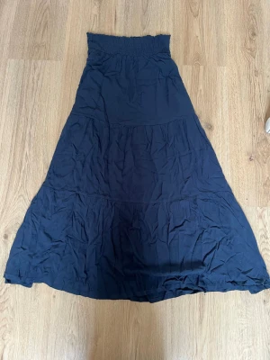 Svart långkjol med smock i midjan - En marinblå långkjol med bred smock i midjan som ger en bekväm och flexibel passform. Kjolen har en lätt utställd form och är sydd i flera paneler för en snygg siluett. Perfekt att styla med en crop top eller en enkel t-shirt.