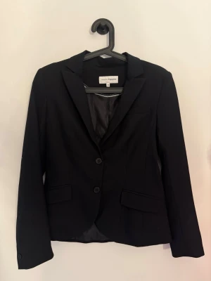 Svart kavaj från KappAhl - Stilren svart kavaj från KappAhl med klassisk krage, två knappar framtill och fickor med lock. Kavajen har figurnära passform och helfodrad insida, perfekt för en snygg och uppklädd look.