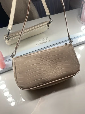 Beige axelremsväska från Even&Odd - Snygg beige handväska från Even&Odd med croco-mönstrad yta och silverfärgade detaljer. Väskan har en avlång form och dragkedja upptill. Perfekt för dig som gillar stilrena accessoarer med lite edge.skriv för fler bilder, pris kan diskuteras!!💗