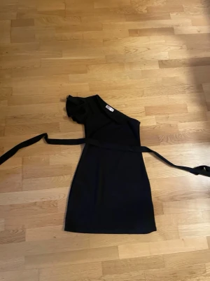 Svart oneshoulderklänning Kilottor - Svart kortklänning från Kilottor med oneshoulder-design och volangdetalj på ärmen. Klänningen har ett långt knytband i midjan som ger en snygg siluett. Perfekt för dig som gillar stilrena och trendiga plagg.