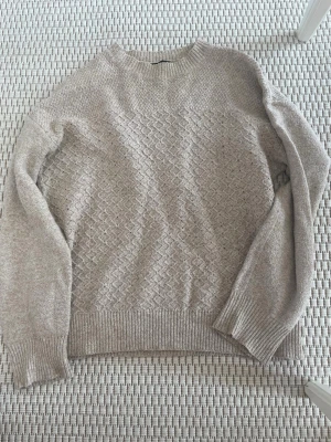 Beige stickad tröja med strukturmönster - Beige stickad tröja med snyggt strukturmönster framtill och på ärmarna. Tröjan har rund halsringning, långa ärmar och ribbade muddar vid ärmslut och nederkant. Perfekt för lager-på-lager och enkel att matcha med jeans eller kjol.