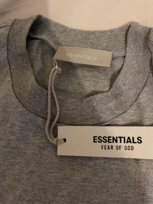 Grå Essentials Fear of God t-shirt - Snygg grå t-shirt från Essentials Fear of God med svart logga tryckt på bröstet. Klassisk rund halsringning och korta ärmar. Perfekt för en clean och stilren streetwear-look. Den är M passar som L