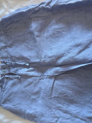 Ljusblå linneshorts från These glory days - Säljer ett par ljusblå shorts i linne med snörning och knapp i midjan. Shortsen har två bakfickor med knappar och elastisk midja för extra komfort. Perfekta för varma dagar och en avslappnad stil.