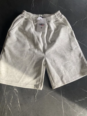 Ljusgrå shorts från Cortiez - M - Säljer ett par Cortiez shorts i ljusgrå/gråmelerad färg. Snygg streetwear-modell med ren look och diskret tryck på benet.  Detaljer: 	•	elastisk midja 	•	sidofickor 	•	bakficka 	•	vit logga/tryck på benet 	•	mjuk och bekväm kvalitet  Skick: bra begagnat skick Färg: ljusgrå / gråmelerad Stil: streetwear / casual  Shortsen säljs enligt bilder. Se gärna alla bilder för egen bedömning av skick och detaljer.  Kan skickas eller hämtas upp enligt överenskommelse.