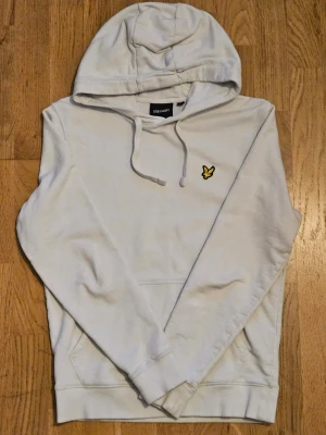Vit hoodie från Lyle & Scott - Snygg vit hoodie från Lyle & Scott med klassisk gul logga på bröstet. Hoodien har dragsko i huvan, magficka och ribbade muddar. Perfekt för en chill och stilren look. Materialet är mjukt och skönt, passar till det mesta.