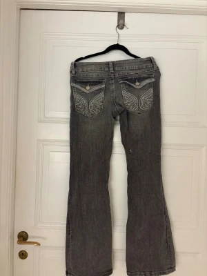 Grå bootcut jeans med broderade fickor - Snygga grå bootcut jeans med broderade bakfickor och knäppning fram. Jeansen har en klassisk femficksmodell och coola detaljer på fickorna som ger extra stil. Perfekta för dig som gillar en avslappnad men trendig look.